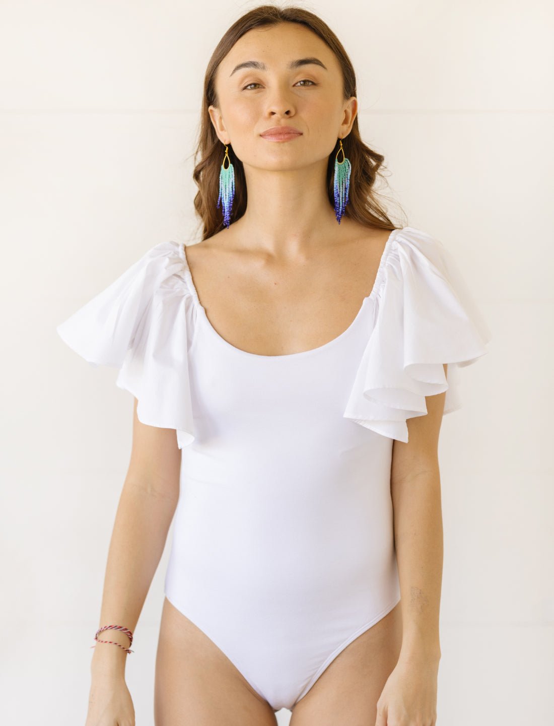 Model wears a white bodysuit with flutter sleeves and a scoop neckline. The bodysuit has ruffled details on the shoulders and a form-fitting silhouette.
