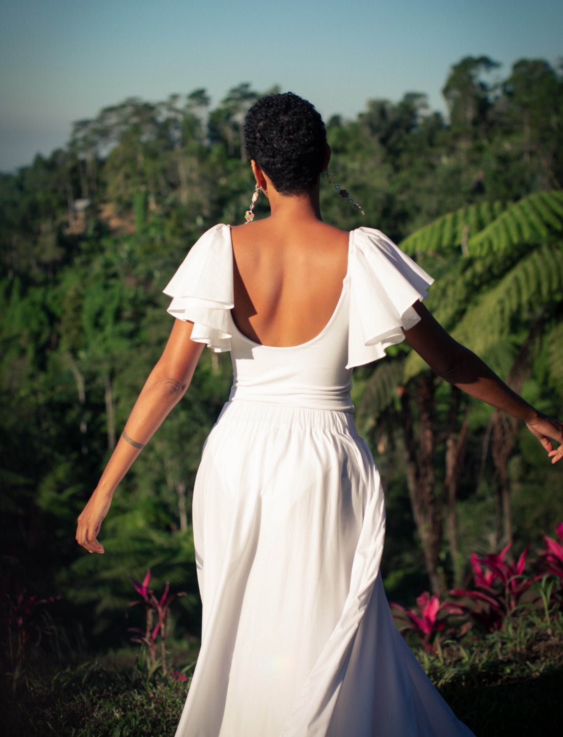 The back view of a woman in a tropical setting. She's dressed in the white Ina Bodysuit with flutter sleeves and a scooped back. The scene is bathed in warm natural light, creating a serene, ethereal mood.