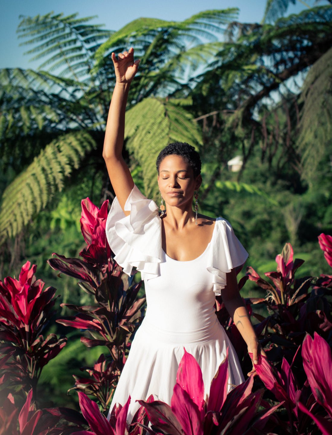 A woman with short dark hair stands in a lush tropical garden with one arm raised in a dancing pose. She is wearing the white Ina Bodysuit with flutter sleeves and a matching skirt. The model has her eyes closed with a serene expression.