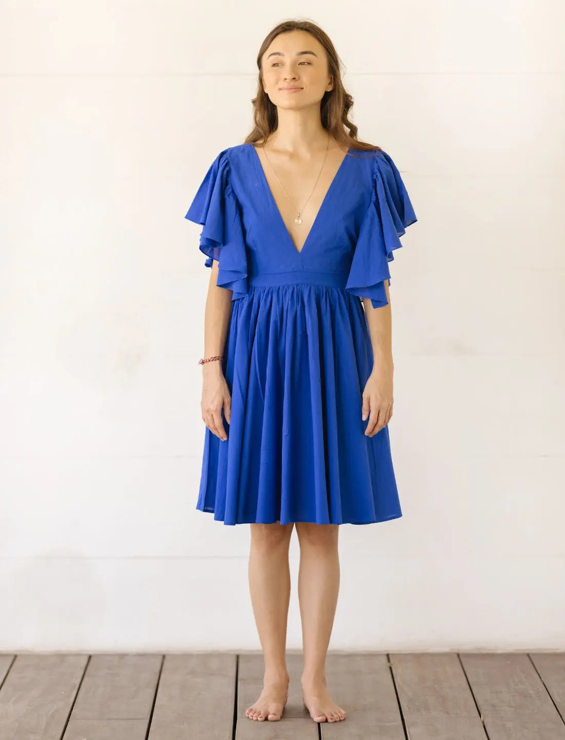 Playful Ina Short Dress in royal blue featuring flutter sleeves, plunging v-neckline and gathered skirt with full silhouette. Ethical fashion, summer cocktail dress, sustainable clothing, handcrafted apparel, wedding guest outfit, bohemian style, vacation dress, beach party attire, artisan made clothing, feminine design, versatile occasion wear photographed against minimalist white background.