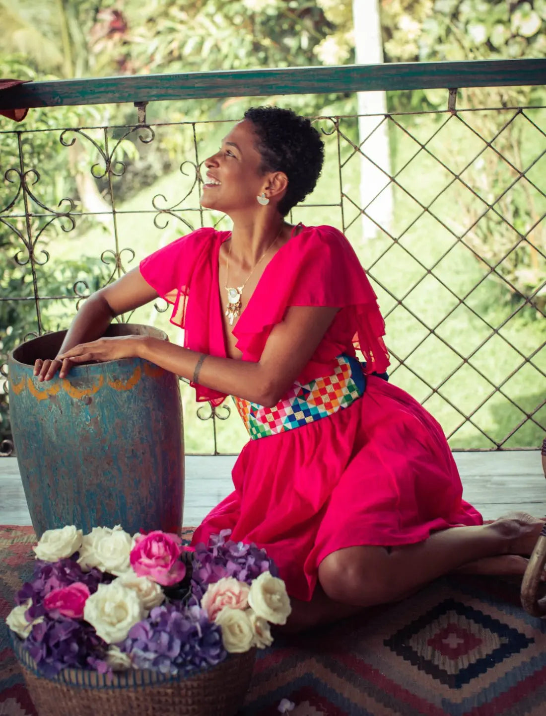 Playful Ina Short Dress in bright fuchsia with Rainbow Wide Belt featuring colorful geometric pattern, v-neckline and flutter sleeves. Bohemian summer fashion, sustainable clothing, ethical apparel, handcrafted artisan belt, tropical resort wear, garden party outfit, vacation dress, statement accessories, Bali-inspired design photographed on balcony with fresh flowers and natural backdrop.