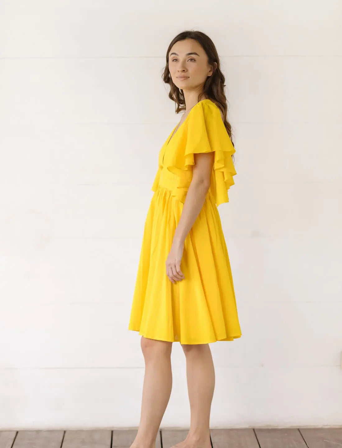 Playful Ina Short Dress in bright yellow with cascading flutter sleeves and gathered skirt, shown from side angle. Summer cocktail dress, ethical fashion, sustainable clothing, wedding guest attire, bohemian style apparel, handcrafted dress, vacation outfit, garden party fashion, feminine silhouette, artisan made clothing, special occasion wear photographed against clean white background.
