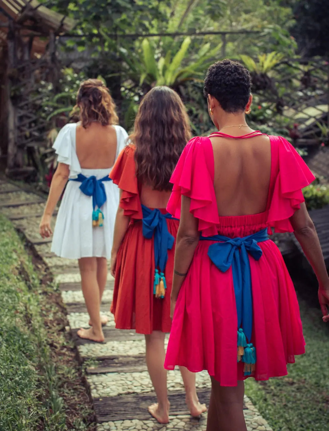 Playful Ina Short Dress collection showcasing open back design in white, terracotta, and fuchsia colors. Bohemian summer fashion, ethical clothing, handcrafted apparel, bridesmaid dresses, destination wedding attire, sustainable fashion, barefoot tropical style, artisan made clothing, beach vacation dresses, open back design, flutter sleeves, tassel details photographed in lush garden setting.