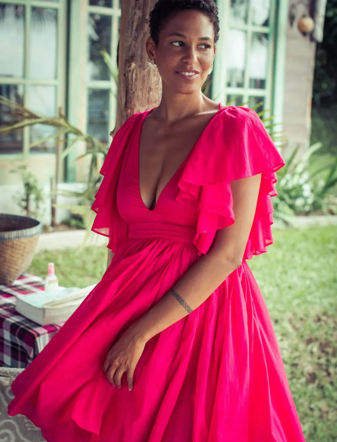 Playful Ina Short Dress in vibrant fuchsia featuring flutter sleeves, plunging v-neckline and flowy gathered skirt. Summer cocktail dress, ethical fashion, sustainable clothing, bohemian style, feminine silhouette, handcrafted apparel, resort wear, tropical vacation outfit, garden party dress, beach wedding guest attire, artisan made clothing photographed in lush outdoor setting.