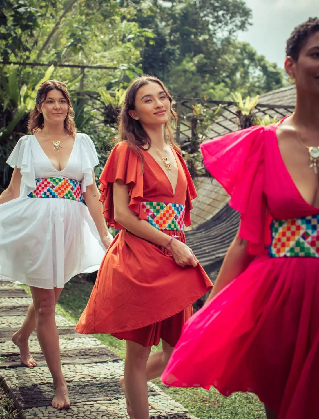 Playful Ina Short Dress collection featuring three vibrant colors - white, terracotta, and fuchsia - each paired with colorful Rainbow Wide Belt. Bohemian bridesmaid dresses, ethical fashion, sustainable clothing, matching wedding party attire, flutter sleeve design, v-neckline, handcrafted apparel, artisan belts, tropical destination wedding, summer garden party outfits, fair trade fashion photographed in lush tropical garden setting.