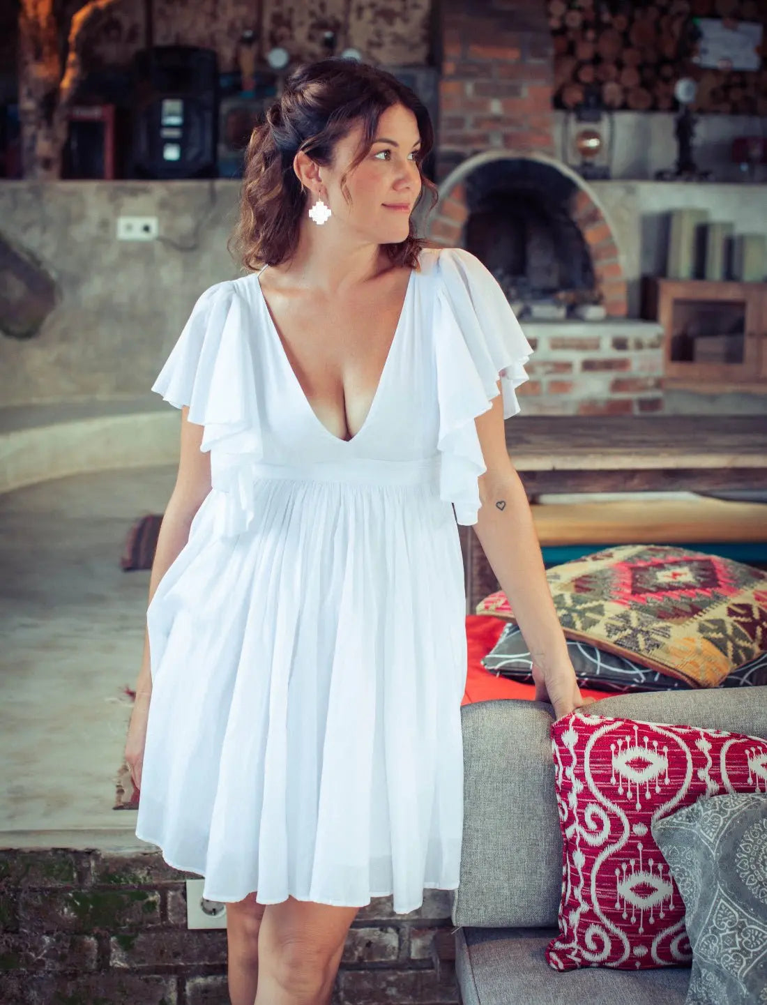Playful Ina Short Dress in crisp white featuring flutter sleeves, plunging v-neckline and flowy gathered skirt. Bohemian summer dress, ethical fashion, handcrafted apparel, bridal shower outfit, honeymoon attire, casual wedding dress, resort wear, statement earrings, sustainable clothing, Bali-inspired design, beach vacation style, feminine silhouette photographed in rustic indoor setting with traditional decor.