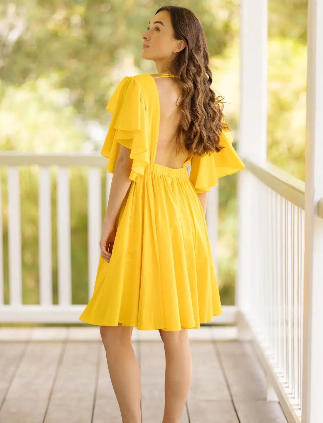 Playful Ina Short Dress in sunny yellow showcasing elegant open back design with flutter sleeves and gathered skirt. Bohemian summer fashion, backless dress, ethical clothing, sustainable apparel, wedding guest outfit, bridal shower dress, beach party attire, handcrafted fashion, vacation wear, garden party dress, outdoor event clothing photographed on wooden porch with natural backdrop.
