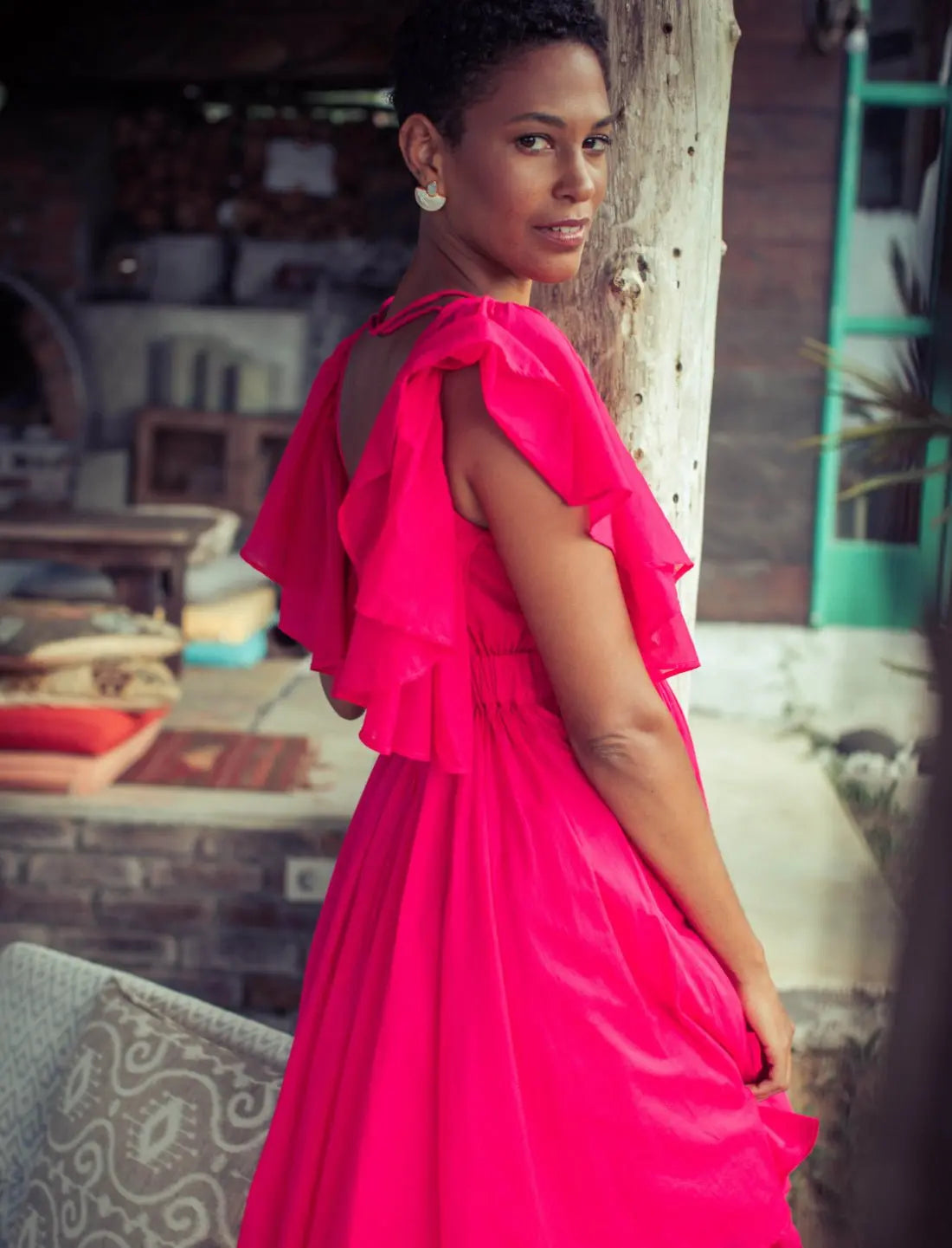 Playful Ina Short Dress in vibrant fuchsia featuring cascading flutter sleeves, open back, and feminine gathered skirt. Bohemian summer fashion, statement cocktail dress, handcrafted ethical clothing, sustainable apparel, tropical vacation outfit, wedding guest attire, romantic date night dress, beach party fashion, artisan made clothing, Bali-inspired design, resort wear photographed in rustic indoor setting.