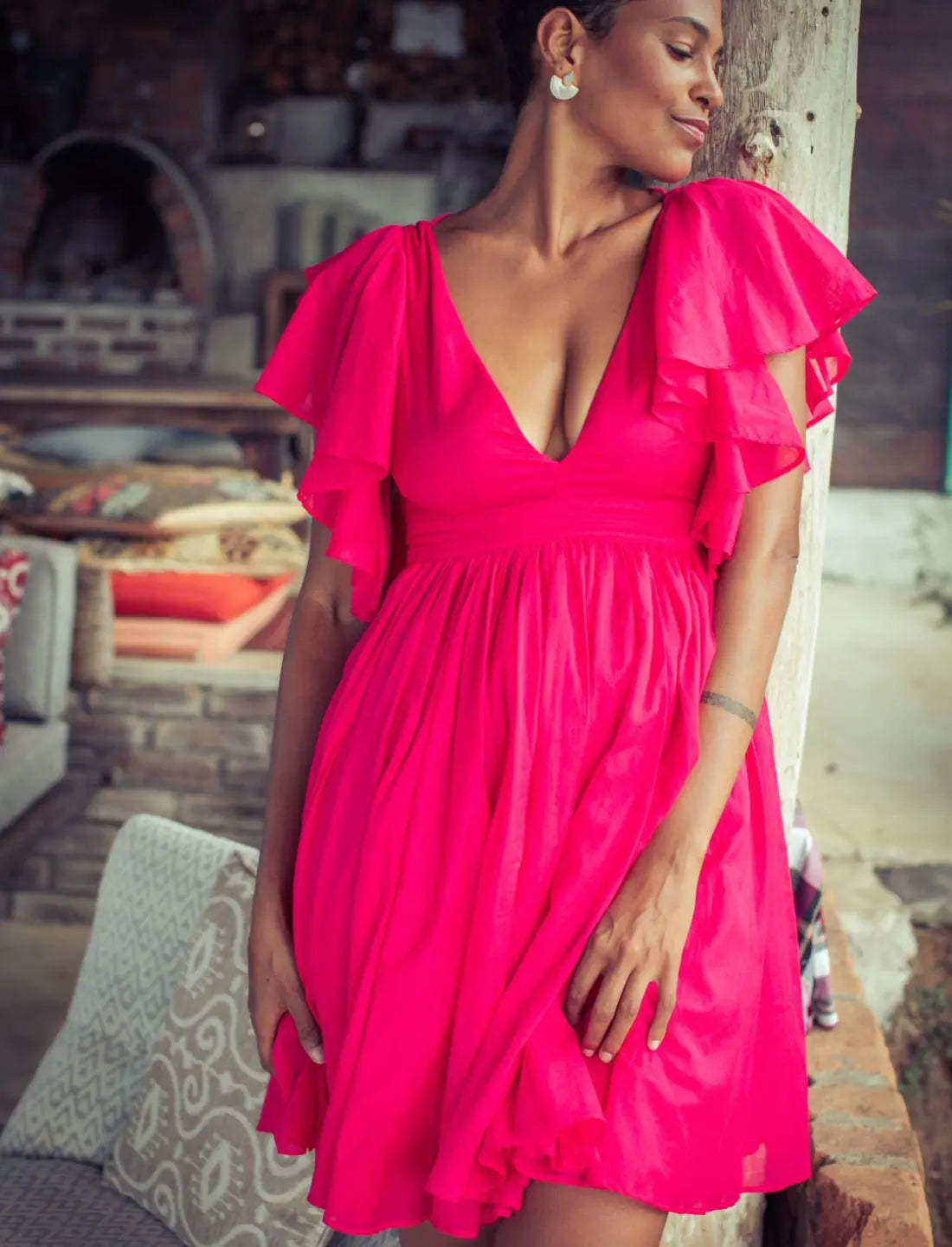 Playful Ina Short Dress in vibrant fuchsia featuring cascading flutter sleeves, deep v-neckline and feminine gathered skirt. Bohemian summer fashion, statement cocktail dress, handcrafted ethical clothing, sustainable apparel, tropical vacation outfit, wedding guest attire, romantic date night dress, beach party fashion, artisan made clothing, Bali-inspired design, resort wear photographed in rustic indoor setting.