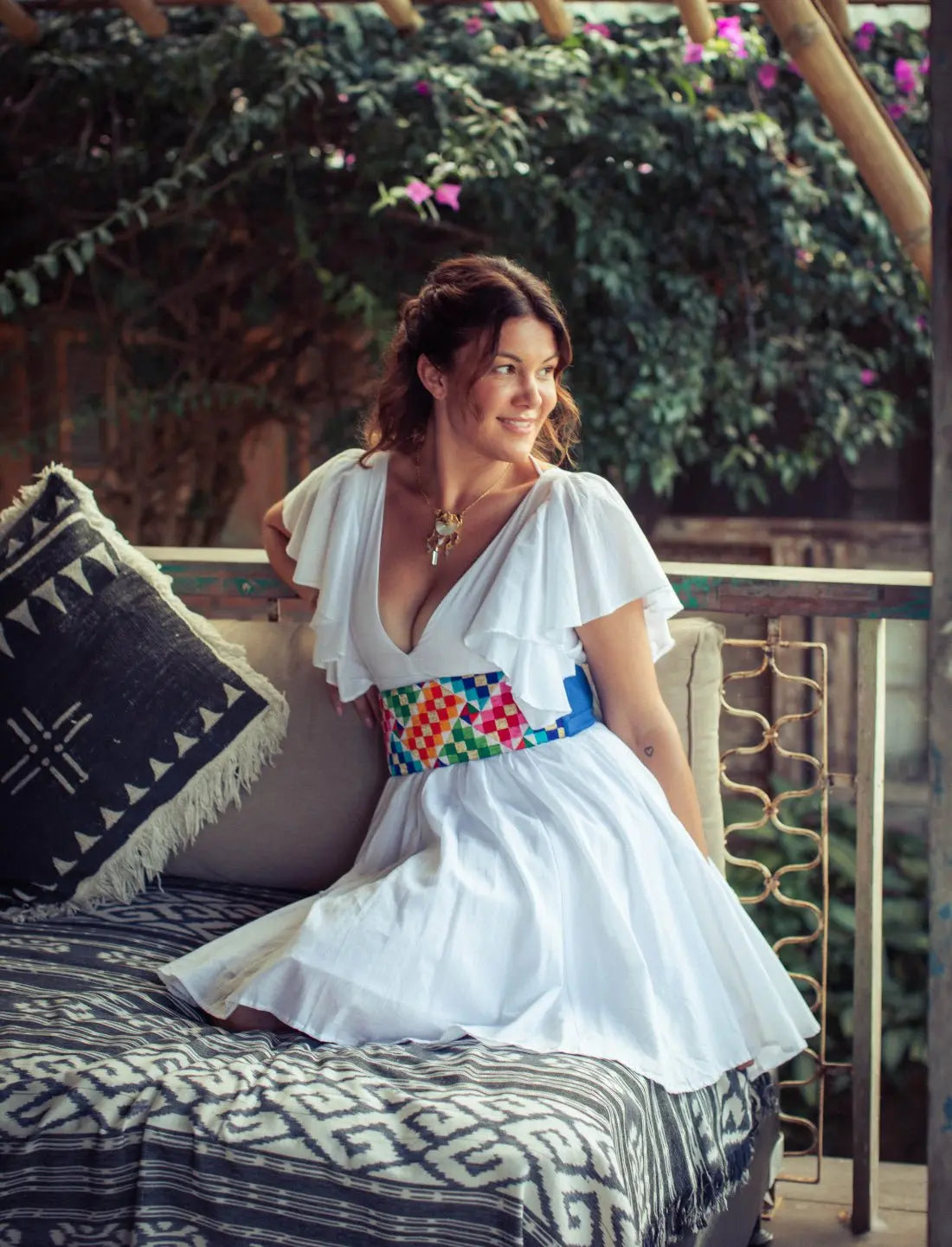 Playful Ina Short Dress in white paired with colorful Rainbow Wide Belt featuring vibrant geometric patterns. Bohemian summer fashion with flutter sleeves, v-neckline, and flowy gathered skirt. Perfect destination wedding guest outfit, ethical clothing, handcrafted fashion, resort wear, vacation dress, artisan accessories, sustainable apparel, Bali-inspired design, tropical style, beach wedding attire photographed in lush garden setting.