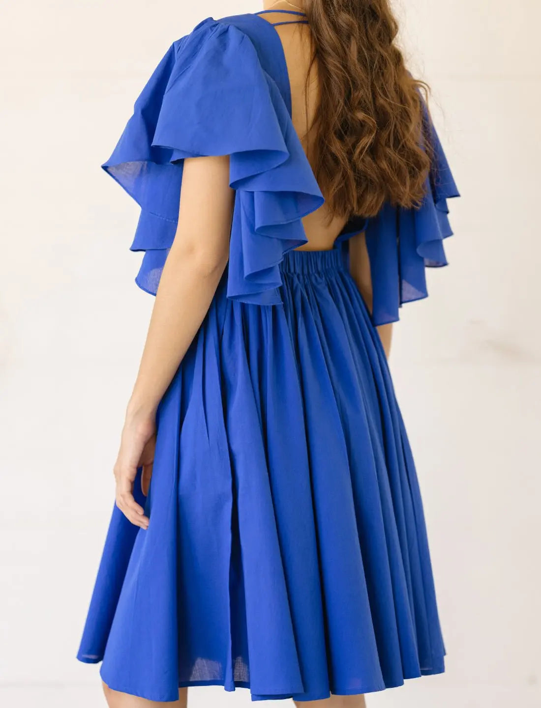 Playful Ina Short Dress in cobalt blue shown from back angle highlighting layered flutter sleeves, open back design with thin straps, and full gathered skirt. Bohemian summer fashion, ethical clothing, sustainable apparel, handmade dress, wedding guest attire, backless design, romantic silhouette, vacation dress, beach party outfit, artisan crafted fashion, special occasion wear photographed against minimalist white background.