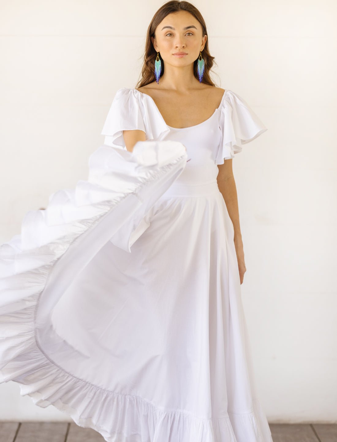 Sacred Hoop of Life Maxi Skirt in pure white with flowing movement, paired with matching flutter sleeve bodysuit top and vibrant beaded earrings. Bohemian wedding dress, ethical fashion, sustainable clothing, beach ceremony gown, simple bridal dress, ruffle hem detail, lightweight fabric motion, handcrafted apparel, minimalist wedding style, artisan made clothing, summer bridal attire photographed against clean white background.