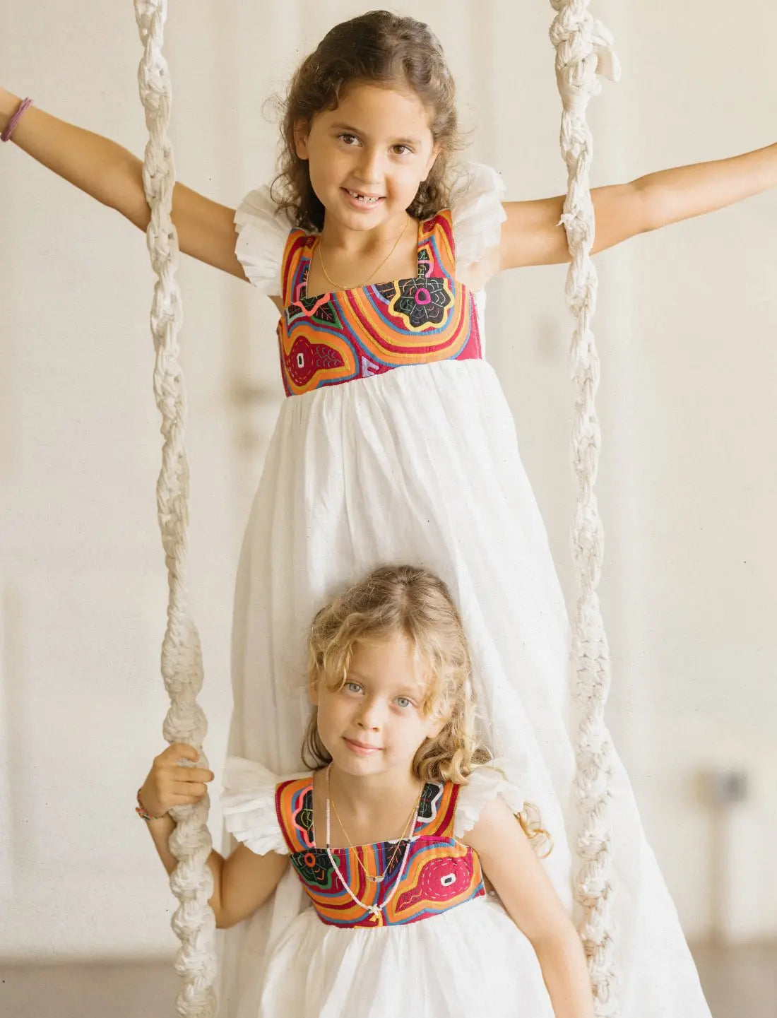 Two young girls wearing matching Yemaya Child Gowns on a rope swing. The older girl stands on the swing with arms extended, while the younger girl sits below. Both gowns feature white linen fabric with vibrant colorful mola embroidery at the bodice showing traditional Kuna designs in red, orange, black, and pink. The dresses showcase the flutter sleeve details and flowing silhouette. Both children are smiling, highlighting how the gowns are both playful and suitable for special occasions.