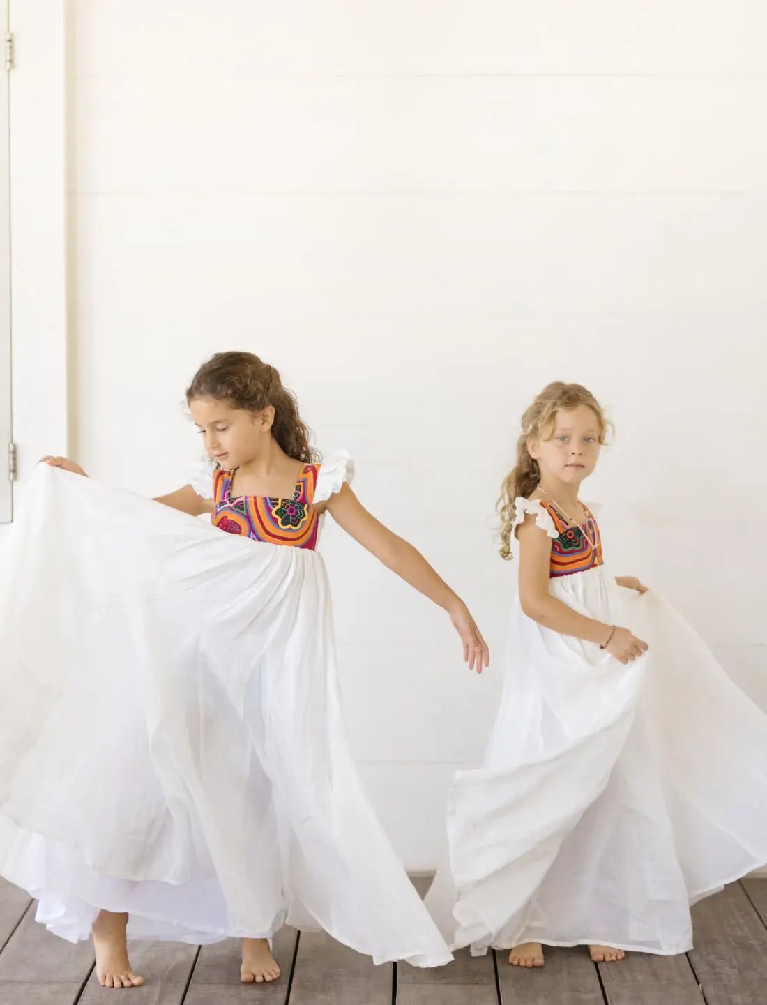 Two young girls wear matching Yemaya Child Gowns in white linen with colorful mola embroidery on the bodice. The girls are standing, mid-twirl. The flowing white dresses feature vibrant orange and red geometric patterns at the neckline, showcasing the traditional Kuna handicraft. The gowns demonstrate their generous fit and playful, twirl-worthy silhouette perfect for children's movement and play.