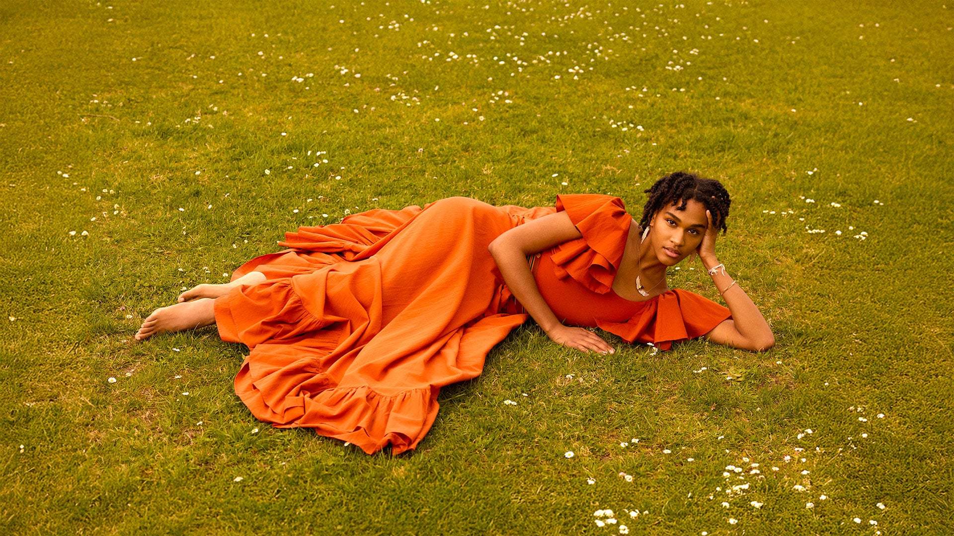 Woman in an orange dress lying on a grassy field with small white flowers.