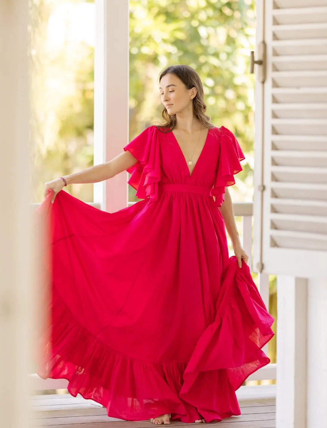 Rich fuchsia Regal Spirit Maxi Dress with flutter sleeves, deep v-neck, and flowy skirt, modeled in a bright home with natural light. Bohemian summer fashion, flowing silhouette, v-neck design, tiered skirt, ethical clothing, resort wear, beach vacation dress, artisan crafted details, sustainable fashion, handwoven accessories, island lifestyle, relaxed luxury, fair trade apparel.