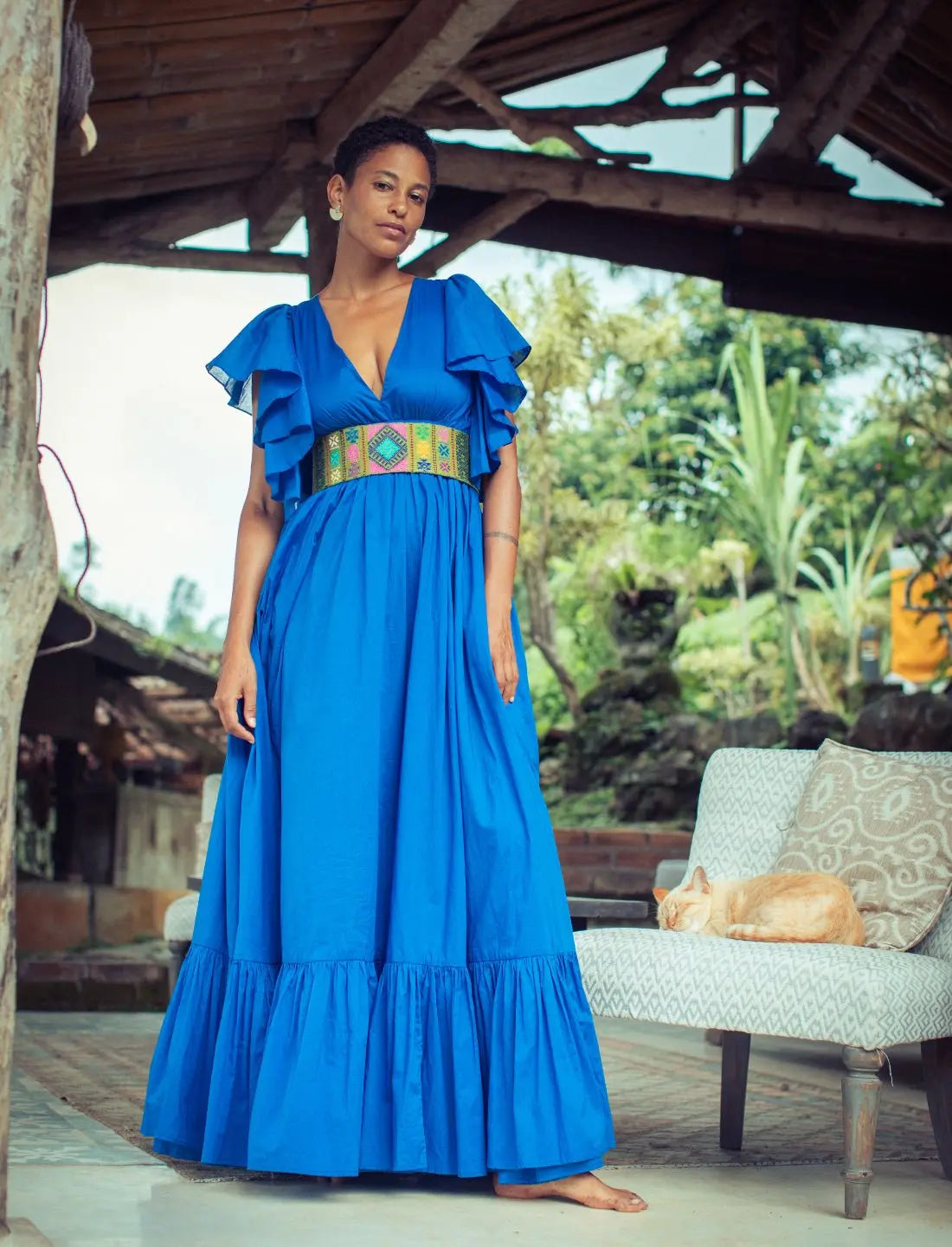 Vibrant royal blue Regal Spirit Maxi Dress with flutter sleeves and a colorful woven belt featuring geometric patterns, modeled in rustic tropical setting with natural light. Bohemian summer fashion, flowing silhouette, v-neck design, tiered skirt, ethical clothing, resort wear, beach vacation dress, artisan crafted details, sustainable fashion, handwoven accessories, island lifestyle, relaxed luxury, fair trade apparel.