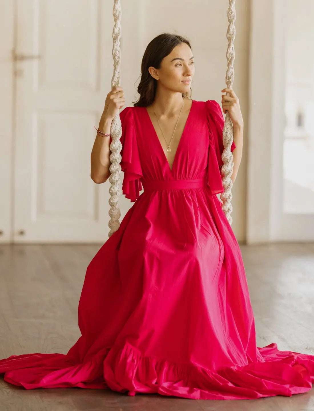 Vibrant fuchsia Regal Spirit Maxi Dress with ruffled flutter sleeves, deep v-neck, and flowy skirt, modeled in a bright home sitting on a swing. Bohemian summer fashion, flowing silhouette, v-neck design, tiered skirt, ethical clothing, resort wear, beach vacation dress, artisan crafted details, sustainable fashion, handwoven accessories, island lifestyle, relaxed luxury, fair trade apparel.