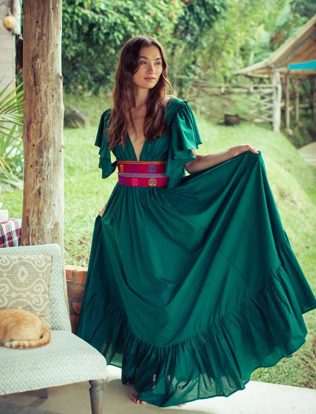 Versatile Spirit Maxi Dress in emerald green with flutter sleeves and colorful embroidered Bhutanese belt, styled on a model in a tropical jungle resort setting. Bohemian fashion, flowy silhouette, ethical clothing, sustainable fashion, forest photoshoot, nature-inspired design, v-neck dress, statement jewelry, eco-friendly apparel, artisan crafted, festival outfit.