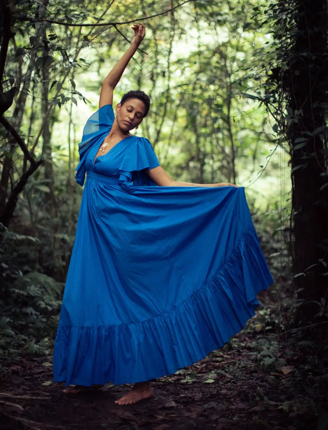 Vibrant royal blue Regal Spirit Maxi Dress with flutter sleeves, and dramatic frill skirt, modeled in a lush tropical setting with natural light. Bohemian summer fashion, flowing silhouette, v-neck design, tiered skirt, ethical clothing, resort wear, beach vacation dress, artisan crafted details, sustainable fashion, handwoven accessories, island lifestyle, relaxed luxury, fair trade apparel.