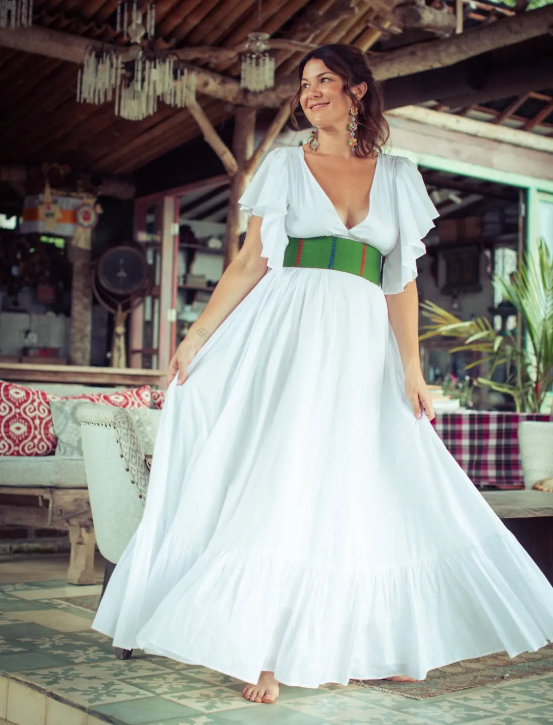 White Regal Spirit Maxi Dress with flutter sleeves and vibrant green tribal-inspired belt, modeled in a rustic tropical beach lounge setting. Bohemian resort wear, flowing white cotton dress, vacation fashion, destination wedding guest outfit, summer beachwear, ethnic waistband detail, v-neck design, statement earrings, sustainable fashion, barefoot elegance, island style clothing.