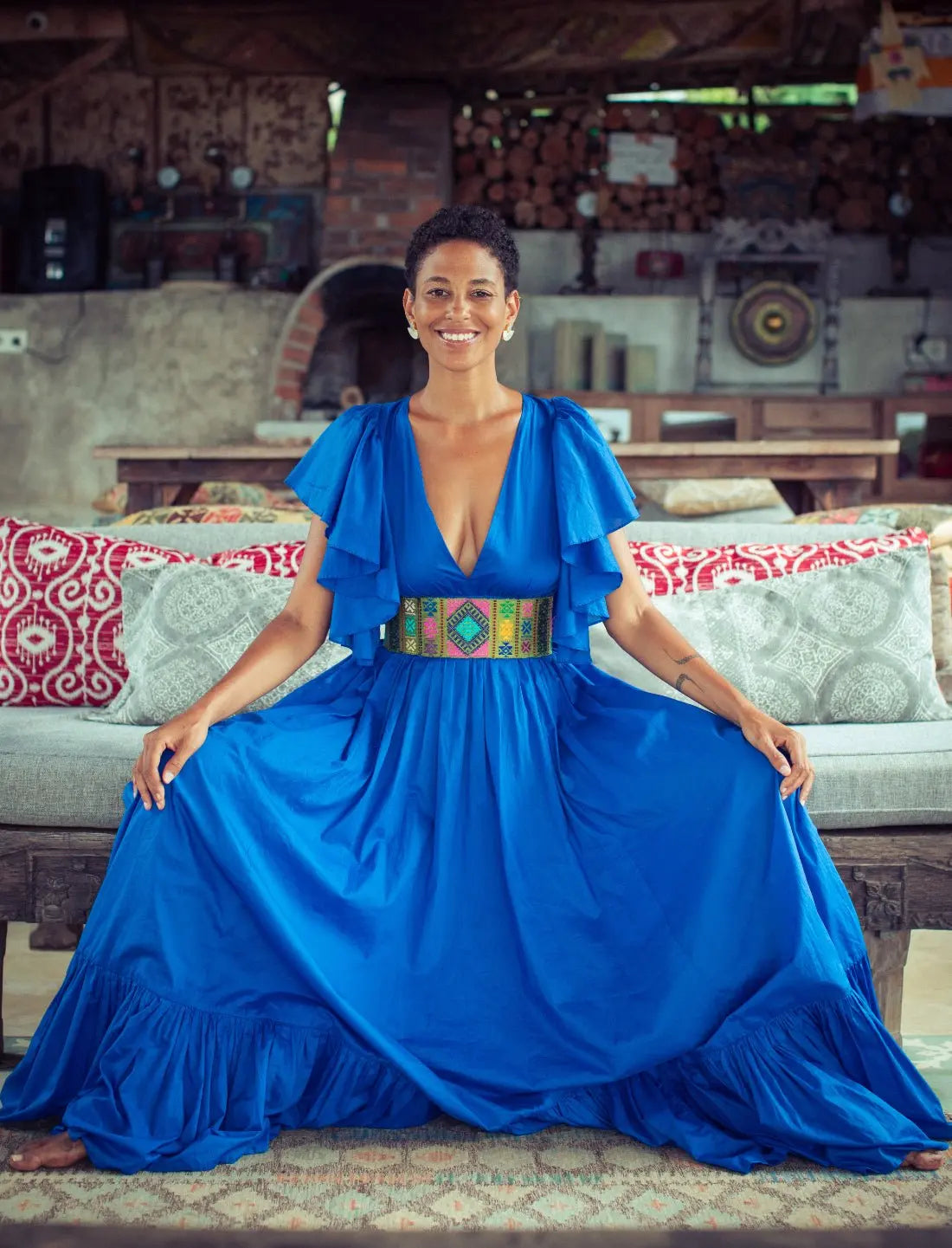 Vibrant royal blue Regal Spirit Maxi Dress with flutter sleeves, and accessorized with a colorful handwoven Bhutanese belt featuring geometric patterns, modeled in rustic tropical setting with natural light. Bohemian summer fashion, flowing silhouette, v-neck design, tiered skirt, ethical clothing, resort wear, beach vacation dress, artisan crafted details, sustainable fashion, handwoven accessories, island lifestyle, relaxed luxury, fair trade apparel.
