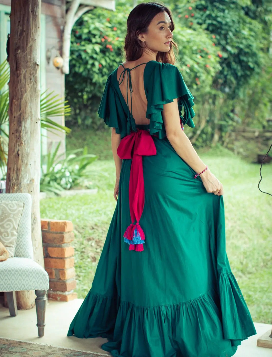 Emerald green backless Regal Spirit Maxi Dress with flutter sleeves and a vibrant red tasseled belt sash, showcasing open back design and ruffle hem. Bohemian tropical fashion, destination wedding guest dress, summer garden party outfit, resort wear, ethical clothing, handcrafted details, feminine silhouette, sustainable fashion, tie-back design, festival dress, boho luxury style photographed in lush garden setting.