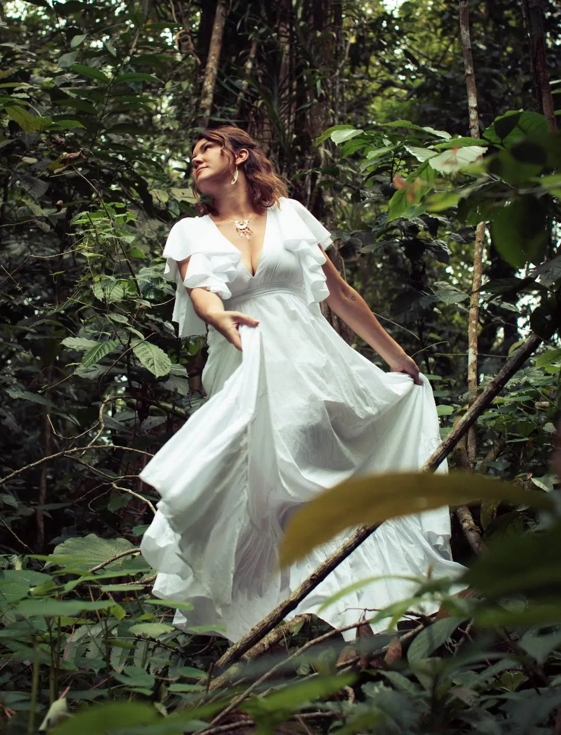 White Regal Spirit Maxi Dress with angelic flutter sleeves, deep v-neck, and flowy skirt, modeled in lush jungle setting. Bohemian summer fashion, flowing silhouette, v-neck design, tiered skirt, ethical clothing, resort wear, beach vacation dress, artisan crafted details, sustainable fashion, handwoven accessories, island lifestyle, relaxed luxury, fair trade apparel.