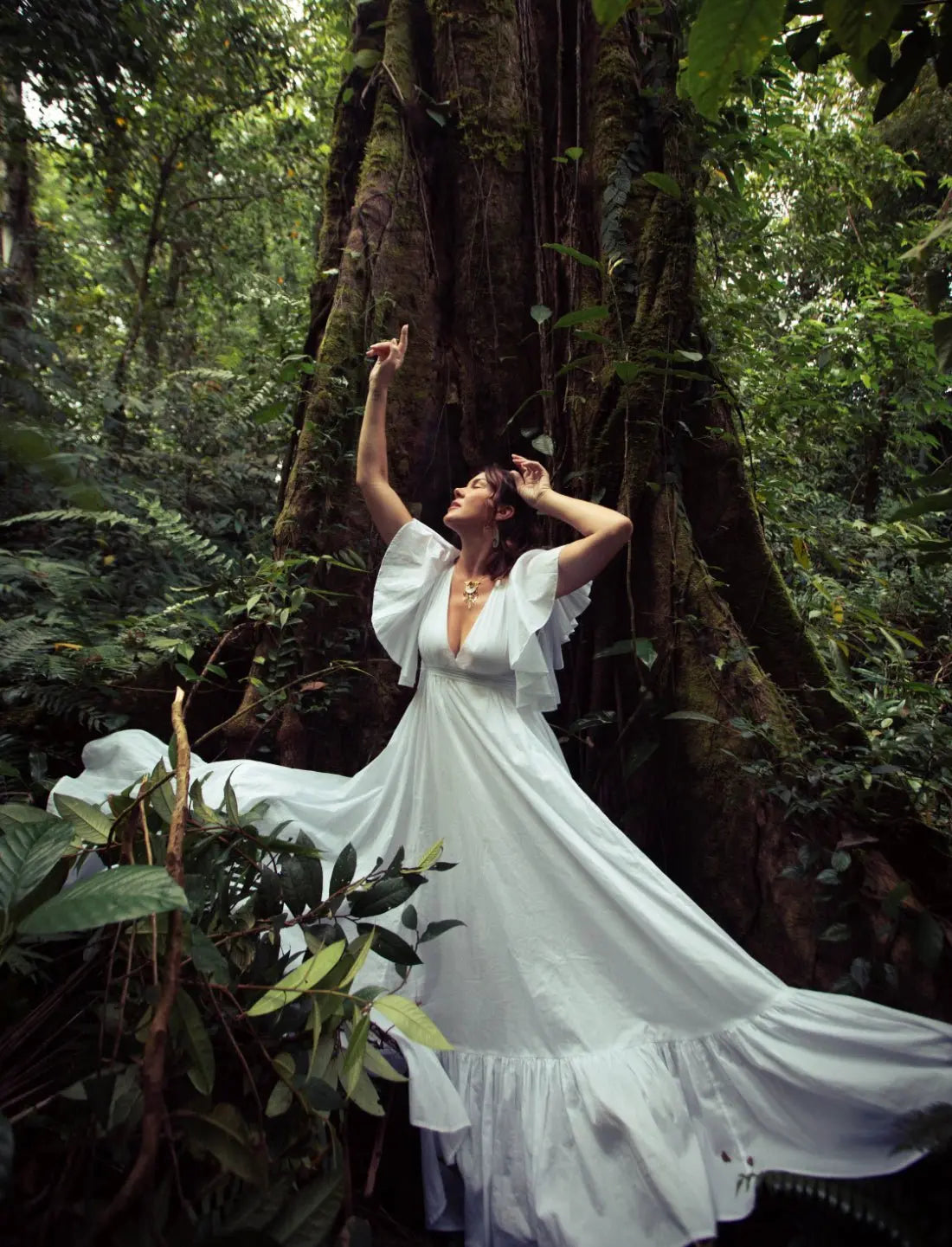Dramatic Regal Spirit Maxi Dress in white with flutter sleeves, deep v-neck, and elegantly flowing skirt, modeled in a lush jungle setting with natural light. Bohemian summer fashion, flowing silhouette, v-neck design, tiered skirt, ethical clothing, resort wear, beach vacation dress, artisan crafted details, sustainable fashion, handwoven accessories, island lifestyle, relaxed luxury, fair trade apparel.