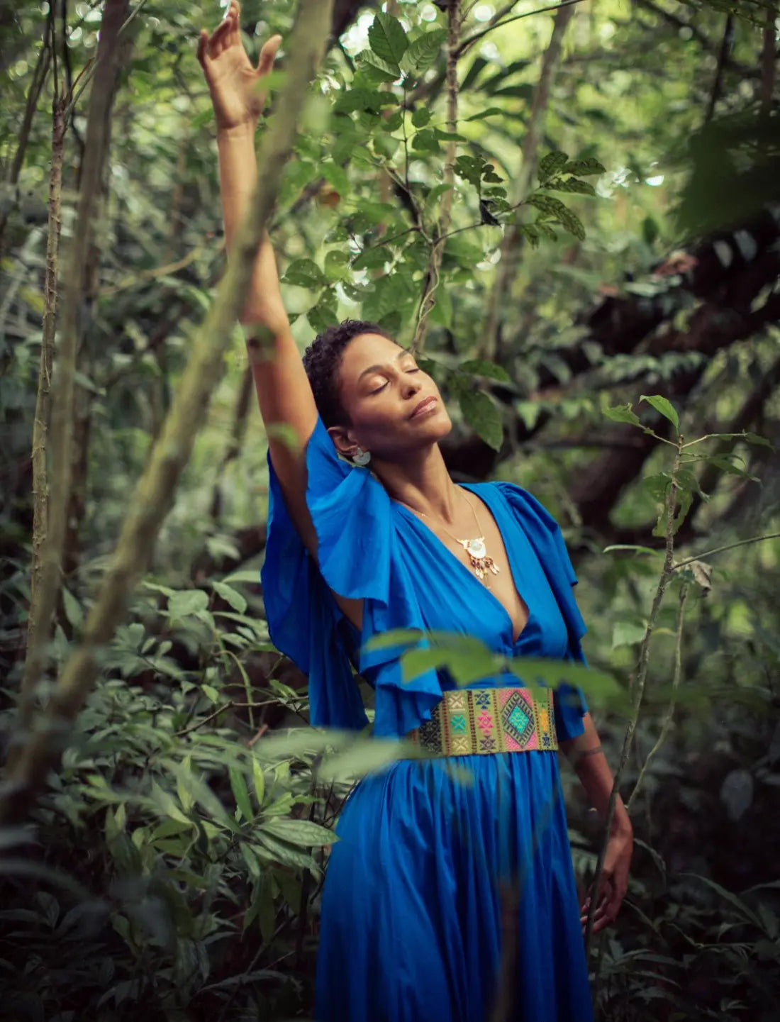 Stunning royal blue Regal Spirit Maxi Dress with flutter sleeves and a colorful woven belt featuring geometric patterns, modeled in a lush jungle setting with natural light. Bohemian summer fashion, flowing silhouette, v-neck design, tiered skirt, ethical clothing, resort wear, beach vacation dress, artisan crafted details, sustainable fashion, handwoven accessories, island lifestyle, relaxed luxury, fair trade apparel.