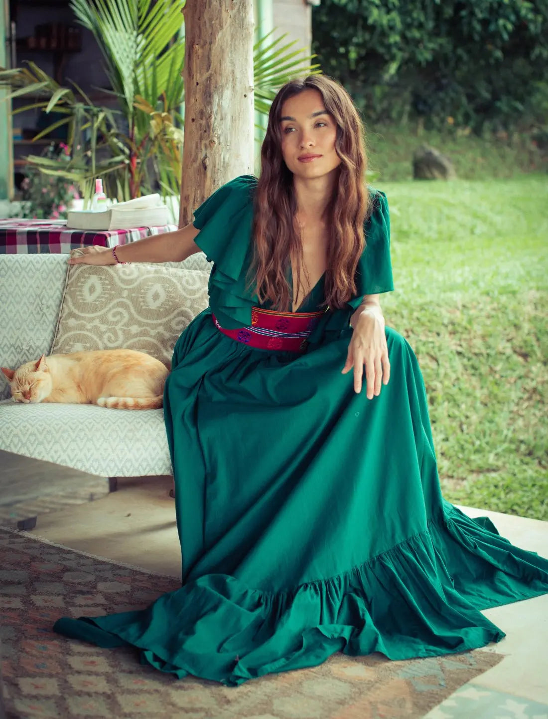 Forest green maxi dress with flutter sleeves and vibrant red Bhutanese belt, showcasing ruffle sleeves and hem. Bohemian tropical fashion, destination wedding guest dress, summer garden party outfit, resort wear, ethical clothing, handcrafted details, feminine silhouette, sustainable fashion, tie-back design, festival dress, boho luxury style photographed in lush garden setting.