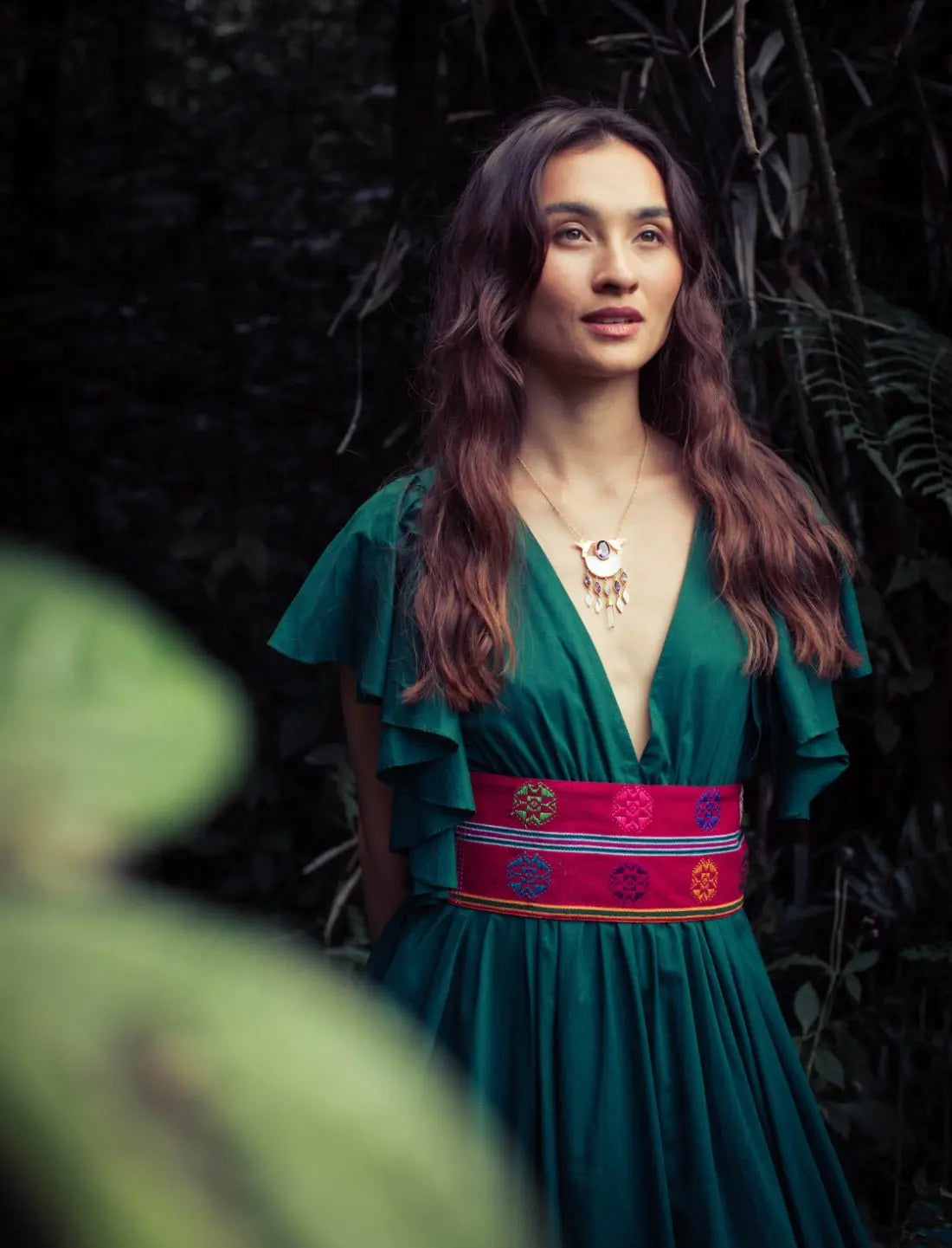 Versatile Regal Spirit Maxi Dress in Forest Green with flutter sleeves and vibrant handwoven belt, showcasing deep v-neckline. Bohemian tropical fashion, destination wedding guest dress, summer garden party outfit, resort wear, ethical clothing, handcrafted details, feminine silhouette, sustainable fashion, tie-back design, festival dress, boho luxury style photographed in lush garden setting.