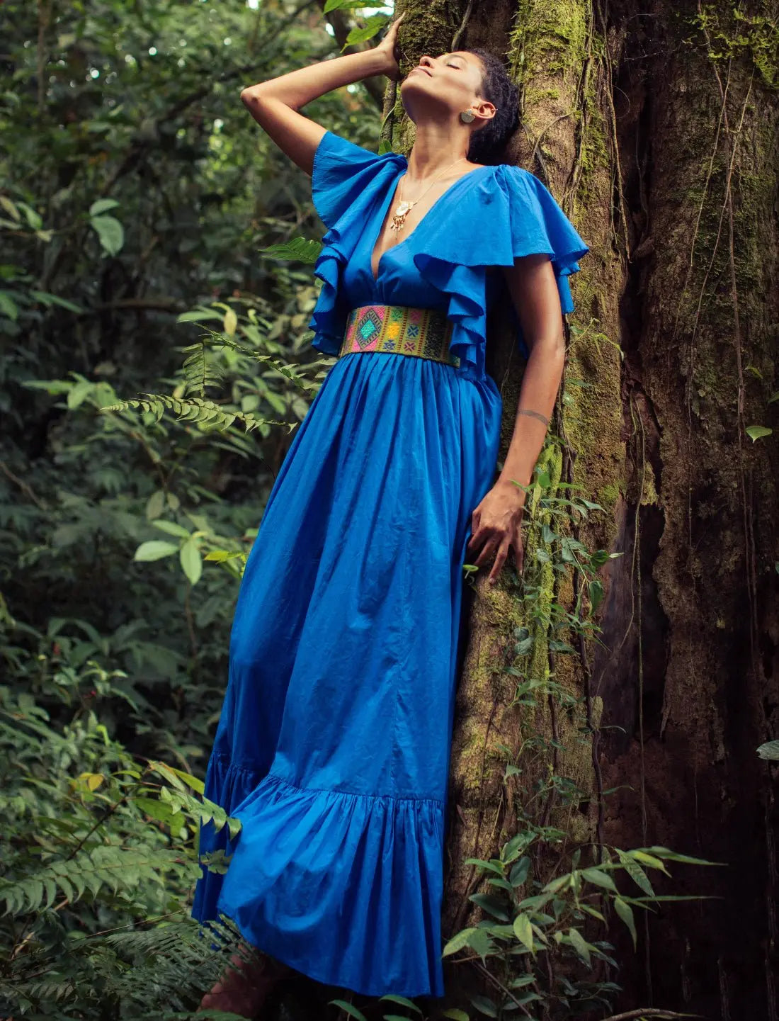 Vibrant royal blue Regal Spirit Maxi Dress with flutter sleeves accessorized with a colorful handwoven Bhutanese belt featuring geometric patterns, modeled in a lush tropical setting with natural light. Bohemian summer fashion, flowing silhouette, v-neck design, tiered skirt, ethical clothing, resort wear, beach vacation dress, artisan crafted details, sustainable fashion, handwoven accessories, island lifestyle, relaxed luxury, fair trade apparel.