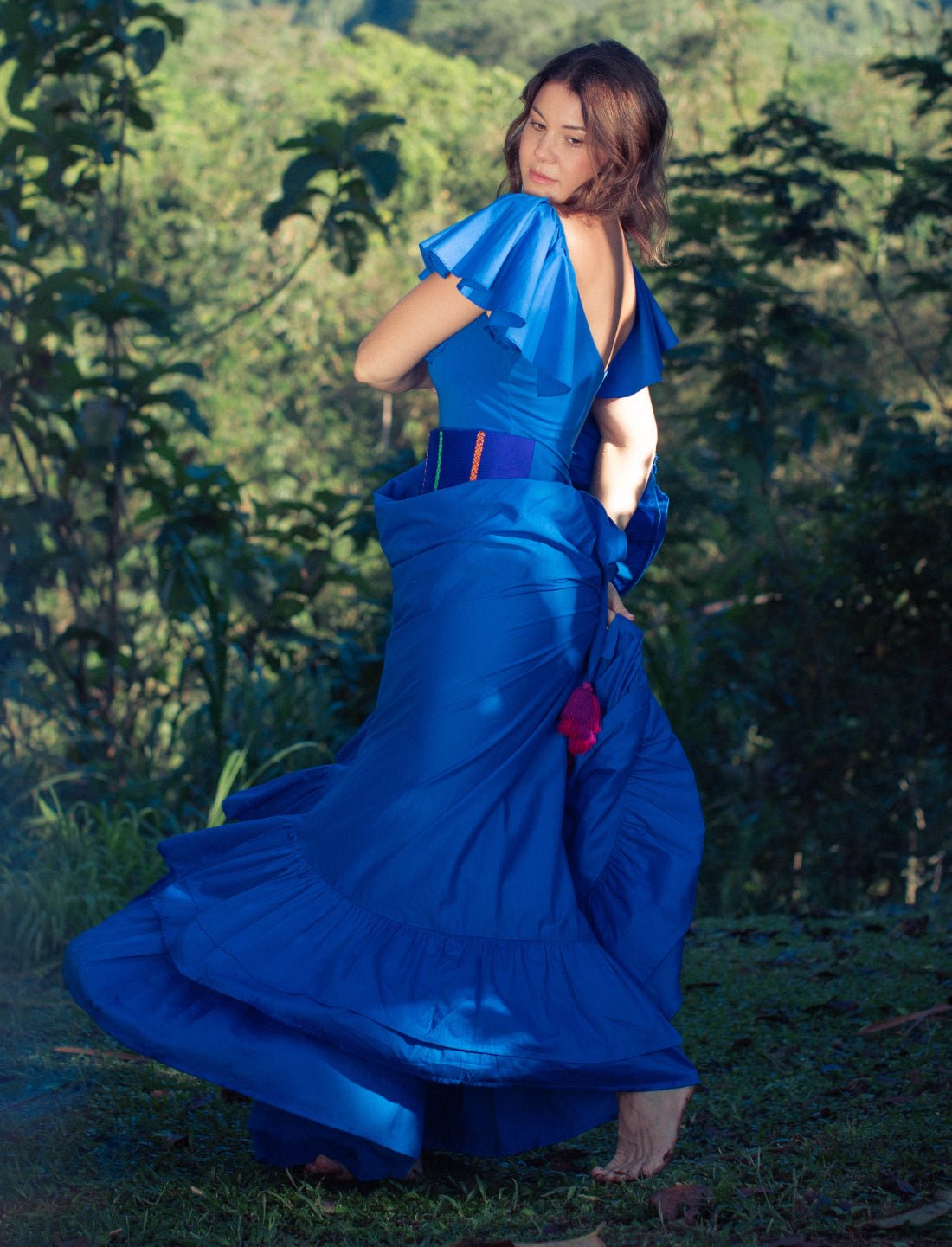 Sacred Hoop of Life Maxi Skirt in royal blue captured mid-twirl with matching flutter sleeve Ina Bodysuit and handwoven Colors of the Cosmos Belt. Bohemian maxi dress, ethical fashion, sustainable clothing, dancing movement, ruffle hem design, barefoot freedom, handcrafted apparel, vibrant blue dress, artisan accessories, outdoor photoshoot, mountain view backdrop, nature-inspired fashion photographed against breathtaking jungle valley landscape.