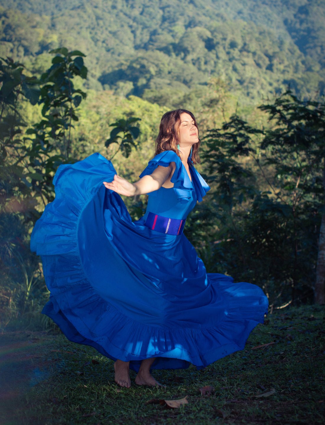 Sacred Hoop of Life Maxi Skirt in royal blue captured mid-twirl with matching flutter sleeve bodysuit and handwoven Colors of the Cosmos Belt. Bohemian maxi dress, ethical fashion, sustainable clothing, dancing movement, ruffle hem design, barefoot freedom, handcrafted apparel, vibrant blue dress, artisan accessories, outdoor photoshoot, mountain view backdrop, nature-inspired fashion photographed against breathtaking jungle valley landscape.