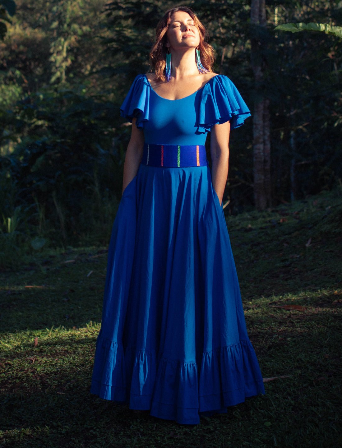 Sacred Hoop of Life Maxi Skirt in royal blue with matching flutter sleeve bodysuit, handwoven Colors of the Cosmos Belt and coordinating tassel earrings. Bohemian maxi dress, ethical fashion, sustainable clothing, serene forest setting, ruffle hem design, sunset golden hour photography, handcrafted apparel, vibrant blue dress, artisan accessories, colorful woven belt, statement earrings, nature-inspired fashion photographed against jungle backdrop.