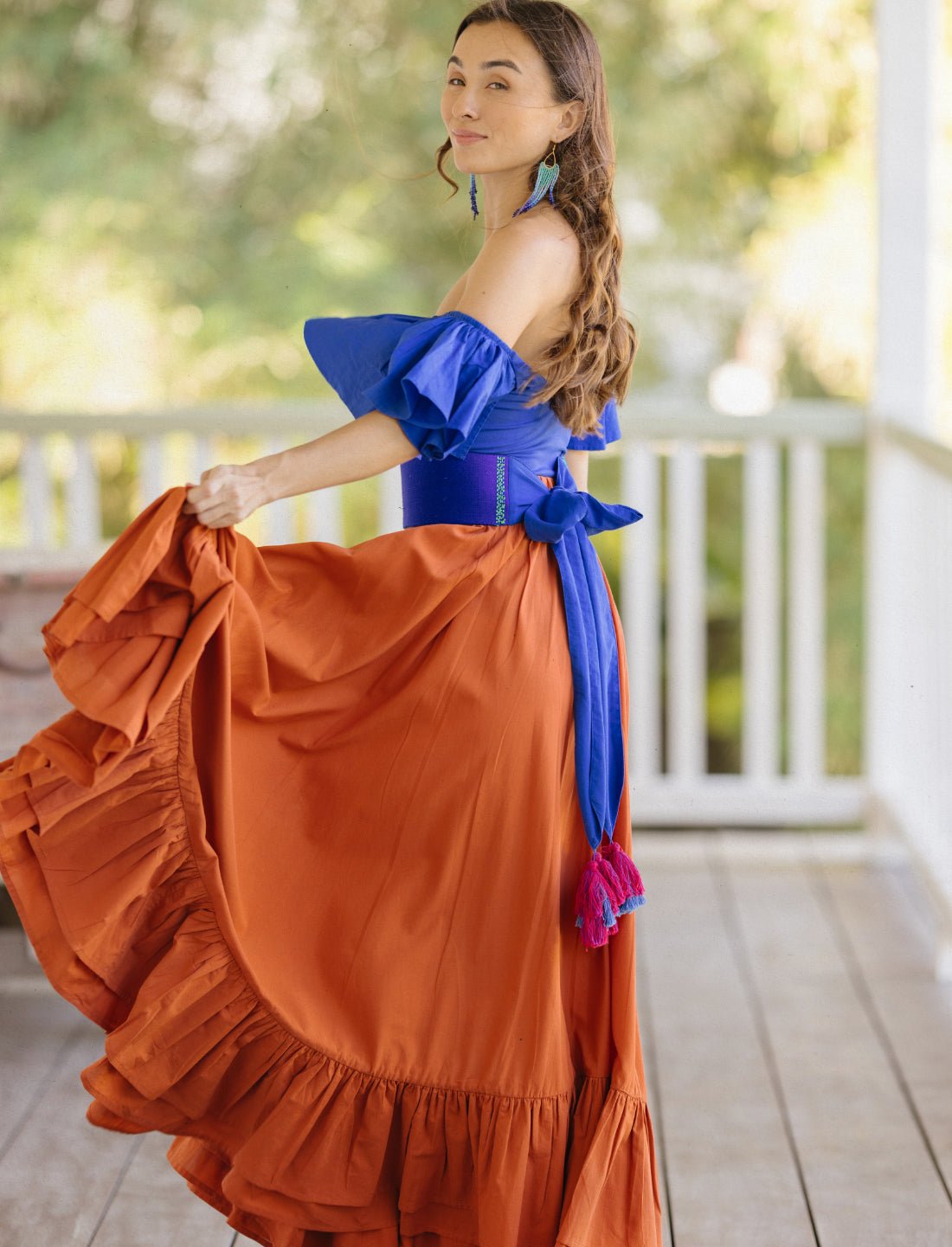 Sacred Hoop of Life Maxi Skirt in terracotta styled with royal blue off-shoulder bodysuit and blue handwoven tassel belt. Bohemian two-piece outfit, ethical fashion, sustainable clothing, colorful boho style, ruffle hem design, summer resort wear, handcrafted apparel, festival outfit, beach vacation clothing, artisan made fashion, statement earrings, flowing silhouette photographed on wooden porch with natural backdrop.