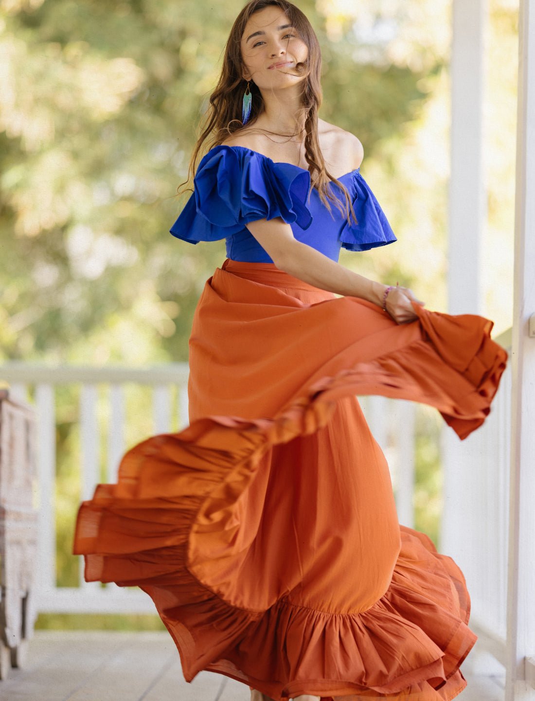 Sacred Hoop of Life Maxi Skirt in terracotta with flowing movement, paired with royal blue off-shoulder bodysuit top and beaded earrings. Bohemian two-piece outfit, ethical fashion, sustainable clothing, ruffle hem design, summer resort wear, festival fashion, beach vacation outfit, handcrafted apparel, danceable silhouette, artisan made clothing, movement photography, boho style photographed on bright wooden porch.