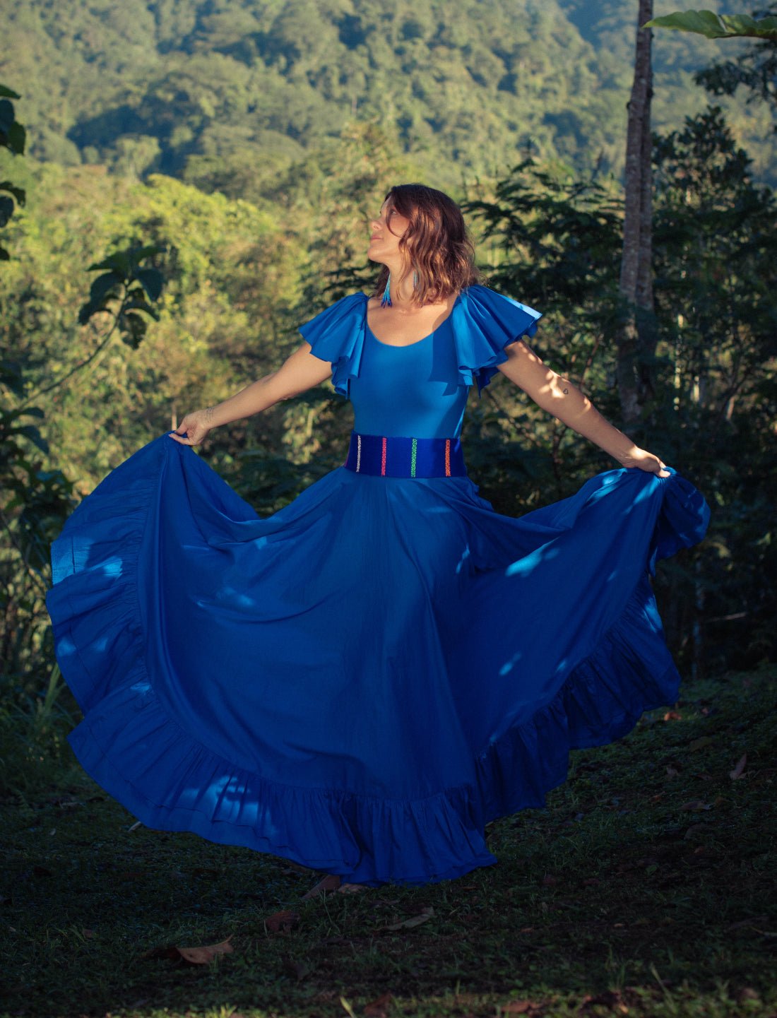 Sacred Hoop of Life Maxi Skirt in royal blue shown in full twirl with matching flutter sleeve top and Colors of the Cosmos Belt. Bohemian maxi dress in motion, ethical fashion, sustainable clothing, mountain backdrop, ruffle hem design, dancing silhouette, handcrafted apparel, vibrant blue dress, artisan accessories, festival fashion, outdoor photoshoot, nature-inspired style photographed against lush jungle mountains.