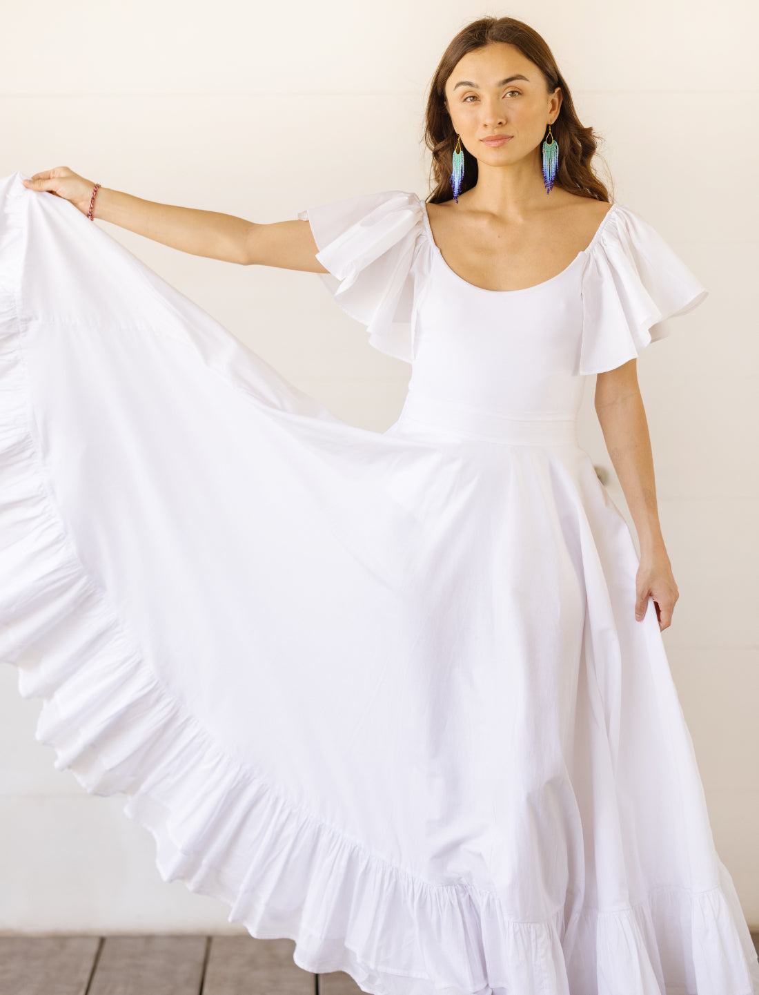 Sacred Hoop of Life Maxi Skirt in pure white shown in movement with matching flutter sleeve bodysuit top and colorful beaded earrings. Bohemian wedding dress, ethical fashion, sustainable clothing, beach ceremony gown, flowing bridal dress, ruffle hem detail, handcrafted apparel, dance-inspired silhouette, artisan made clothing, minimalist wedding style, summer bridal attire photographed against clean white background.