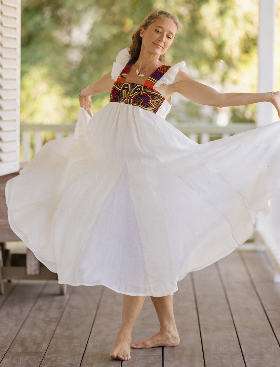 Model wearing the Yemaya Embroidery Adult Gown in white linen with colorful geometric mola embroidery at the bodice. This particular mola pattern includes two colorful pelicans on the front. The model is holding the gown in a majestic pose, the skirt pulled out by her sides into a graceful silhouette. The flowing gown features flutter sleeves and the skirt is made up of a large amount of fabric for maximum flow.
